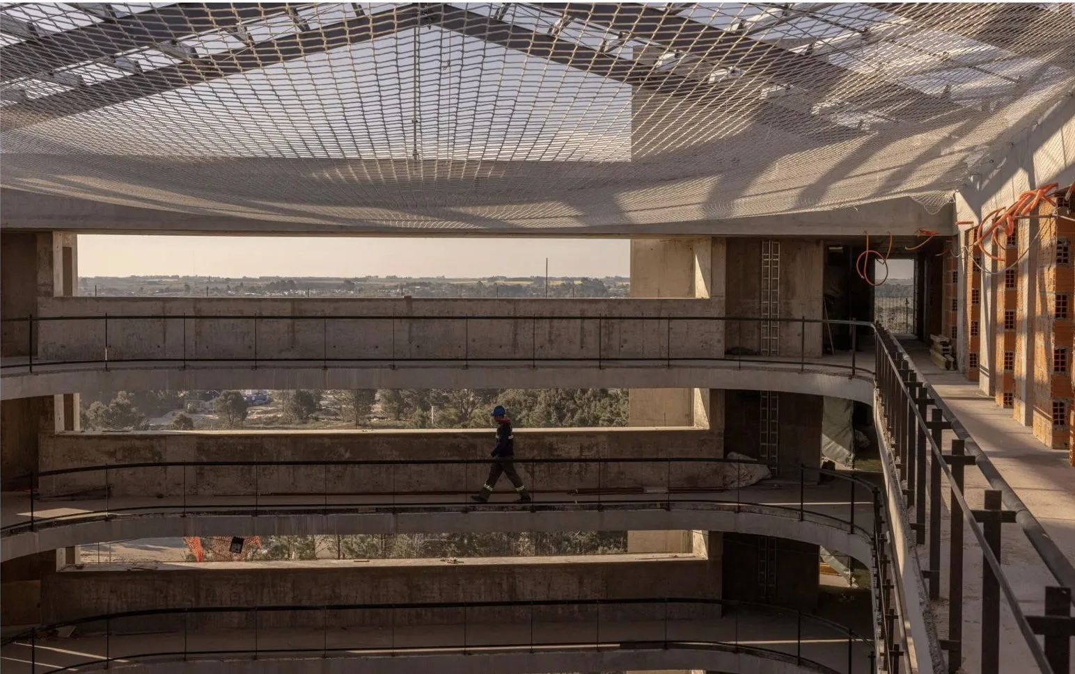 Worker crossing an open concrete atrium beneath a safety net, with fields and trees beyond.